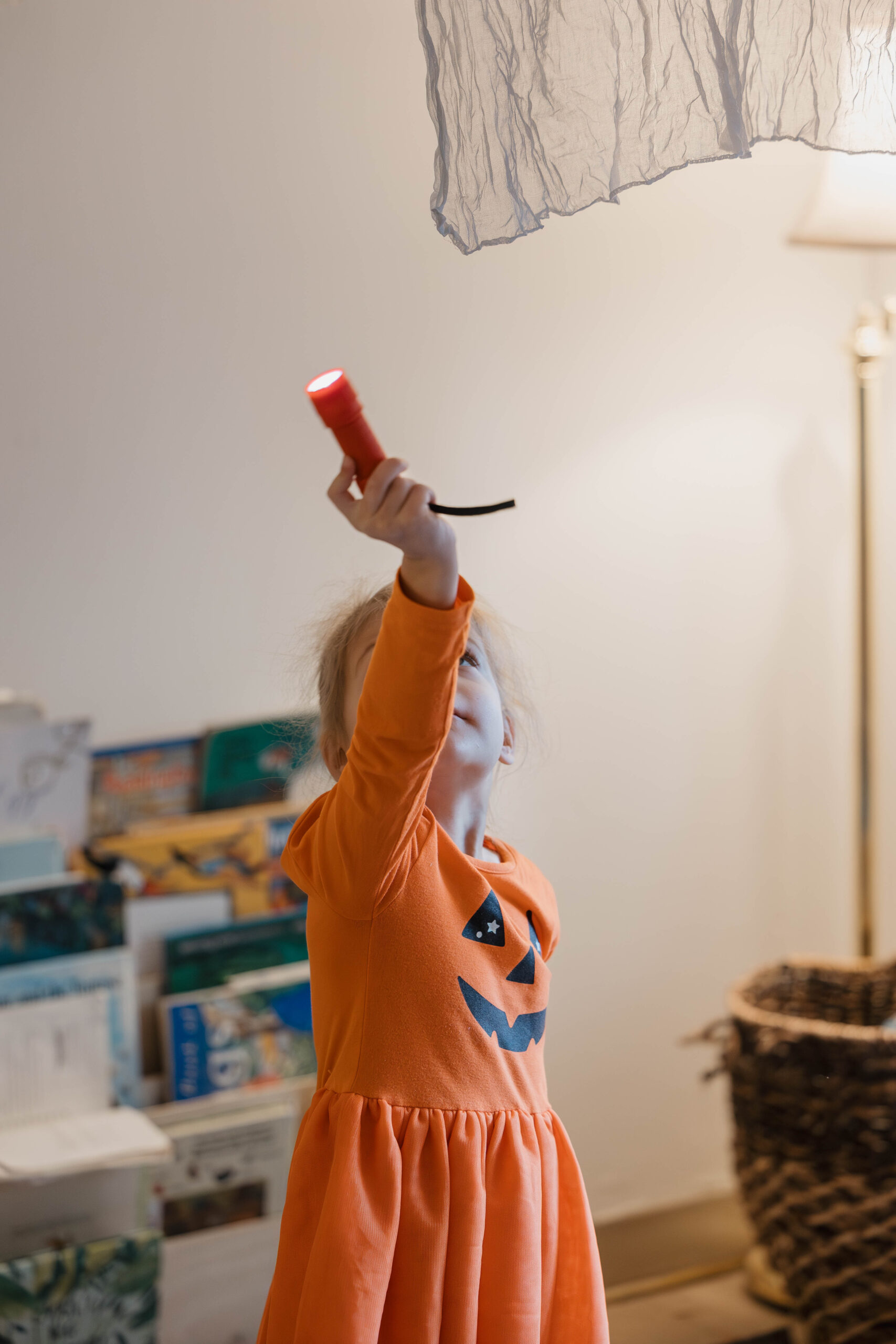 A girl in an orange dress reaching up toward a paper sculpture hanging in the classroom
